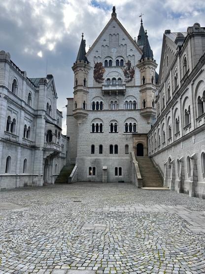 Ein letzter Pflastersteinplatz, links und rechts durch graue Schlossmauern eingegrenzt. Man blickt auf ein Schlossgebäude mit Freitreppe rechts und kleinerer links. Das Gebäude hat links und rechts auch zwei kleine Erker oben.