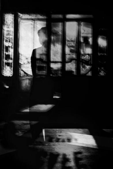 Silhouette einer Figur, die vor einem Hintergrund aus verzierten Fensterrahmen steht, mit abstrakten Schatten- und Lichtmustern in Schwarz und Weiß. (KI generierte Beschreibung)