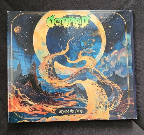 A CD album on dark background. It's very colourful and depicts a fantastic riverscape at night. There are multiple stars in the dark blue sky and two moons, the bigger of which is in the middle and forms an "O". There are dark rocky formations and a couple of trees to the left and right. Between them is a wide river in orange, red and yellow. The fluid in the river has bubble looking things embedded in it. A few tendrils of the liquid are floating up in the middle, curling into the sky through the big moon. The band name "Octoploid" is recognisable in it's light green logo under the upper edge, album name "Beyond the Aeons" is in a small polygonal panel above the lower edge.