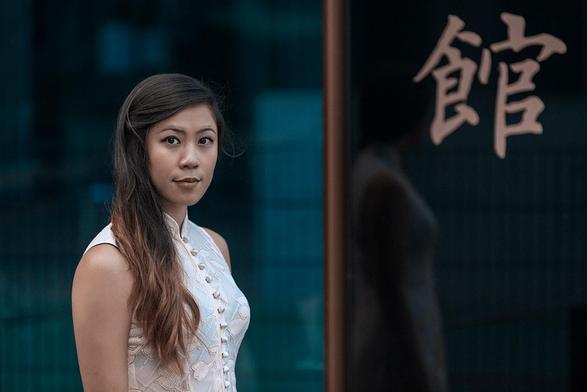 Asiatisch aussehende Frau steht in einem traditionellen Kleid vor einer Scheibe und schaut den Betrachter an. In der Scheibe ist ihre Spiegelung zu sehen, sowie ein asiatisches Schriftzeichen.