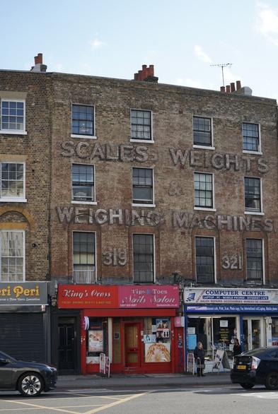 Photo d’une vieille façade d’immeuble en brique au bord d’une rue passante portant un texte en lettres peintes un peu passées : SCALES - WEIGHTS - WEIGHING MACHINES - 319 -321. Le rez de chaussée est occupé par des commerces aux enseignes colorées (esthéticienne, café Internet, etc.)