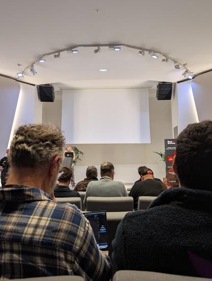 La photo de la scène d'un auditorium. Elle est prise depuis un siège du public ; la scène est vue entre deux dos de membre du public.

L'auditorium est de taille moyenne, blanc, très moderne.