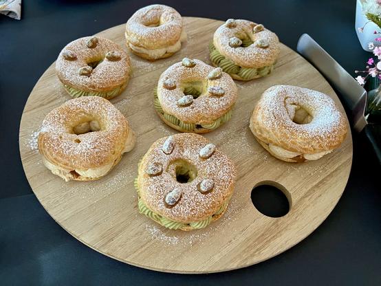 Seven choux pastry ring donuts filled with pistachio whipped buttercream on a wooden board, sprinkled with powdered sugar and topped candied  pistachios