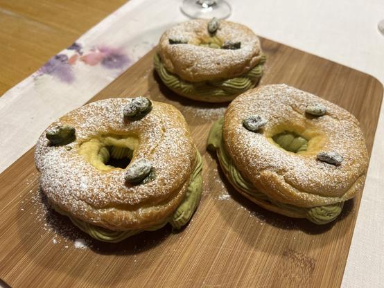Three choux pastry ring donuts filled with pistachio whipped buttercream on a wooden board, sprinkled with powdered sugar and topped candied  pistachios