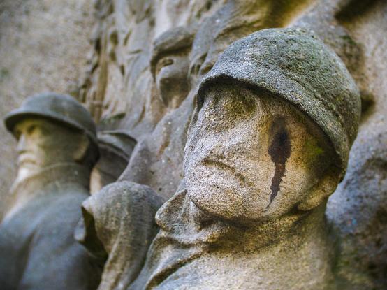 A close-up shot of a stone monument depicting a group of soldiers. The soldiers are wearing helmets and have stern facial expressions. The stone is weathered and has some dark streaks running down the faces of the soldiers.