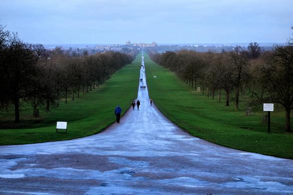 A distant shot of a long, straight path leading to a castle in the distance. The path, wet and reflective, stretches through a park-like setting with bare trees lining both sides. People are walking along the path. The sky is overcast and grey. The perspective suggests the photo was taken from a slightly elevated position.