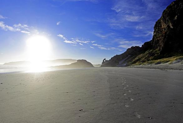 Photo shows a morning at the beach. The sky is blue with scattered clouds. The rising sun washes out the left side of the photo as a visual source of light.  The lower half of the photo shows light grey sand patterned by the freshly recessed water tide. Only a set of foot steps to the right side breaks the natural appearance. In the background there are a stony and hillside landscape dramatically in shadows and foggy mist. Sharp stone formations including a small heart shaped keyhole is visible just off the centre opening between the rocks which makes the entrance to the beach.