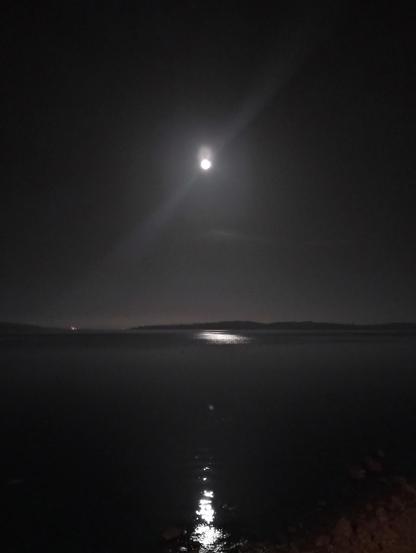 Imagen nocturna de un cuerpo de agua oscuro y tranquilo, la bahia de Ancud, bajo una luna llena brillante. La luna, situada en el centro superior de la imagen, ilumina ligeramente la superficie del agua, creando un sendero de luz reflejada. En la distancia, una línea oscura y levemente visible representa la orilla opuesta. El cielo es predominantemente negro, contrastando con el brillo de la luna. La escena transmite serenidad y calma.