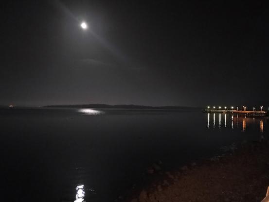 Una vista nocturna de un cuerpo de agua, la bahia de Ancud, tranquilo bajo una luna brillante. En la distancia, se puede ver una línea de costa oscura y un muelle iluminado con luces tenues que se reflejan en el agua. El cielo es oscuro y la luna es el elemento más brillante de la escena. La atmósfera es serena y pacífica.