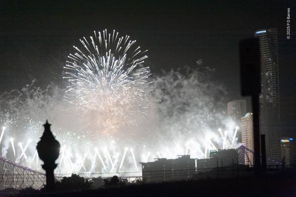 Fountains of criss-crossing white fire shoot up from the Storey Bridge, while overhead concentric spherical white airbursts light up buildings and drifting smoke.