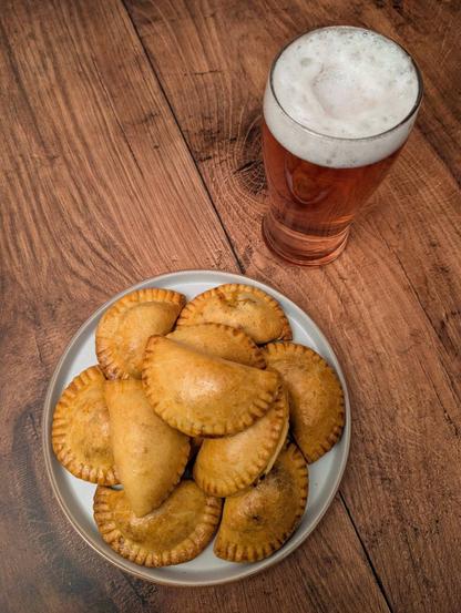 A photo of a plate with a pile of small semi-circle pie bites on it next to a pint glass with a drink in it. The bites are a shiny golden-brown, with little fork ridges along their edges. The drink is a light purple color with a white beer head. They sit on a brown wooden background.