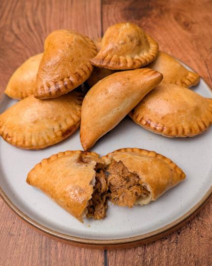 A photo of a plate with small semi-circle pie bites on it, with one at the front pulled open to expose its filling. The bites are a shiny golden-brown, with little fork ridges along their edges. The open one shows a red/brown pulled pork inside, and a nice pasty pastry with some layers but also enough strength to it not to crumble when pulled apart. The plate sits on a brown wooden background.
