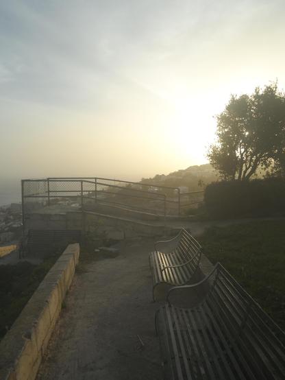 Looking from theGarghur side of the hill towards Madliena hill, the sun rise has painted the sky a golden hue, pushing everything else into silhouette. In the foreground two green benches reflect the morning sunlight