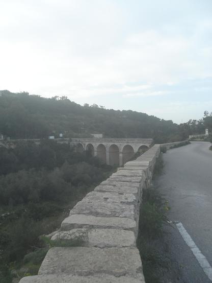 Image is of the bridge that separates Garghur from Madliena and Sweiqi, known as the Victoria lines, it is part of the 12km of fortifications built by the British in 1897 to protect the island. The Bridge is an aqueduct over the valley offering a safe route across for vehicles between the hills that separate Madliena, Sweiqi and Garghur. The hills are lined with tree, and the gorge below is filled with scrub and trees. The sky is powdery blue with whispy white clouds