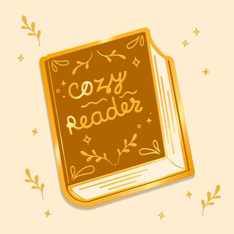 An image with on it the depiction of a golden enamel pin. The pin is a thick soft brown hardcover book. The front cover is decorated with ornate details such as vines and stars, all surrounding the words "Cozy reader".