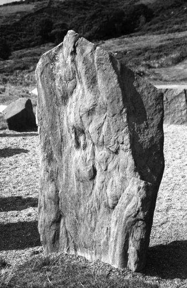 A black and white image of one of the two portal stones that form the Ciorcal Cloch an Droma Bhig (Dromberg Stone Circle). The sun is shining and the stones are casting a shadow, left to right. The stone is largely rectangular but the top has been shaped into a scalene triangle with left hand side being steeper and shorter than the right. The natural weathered face of the stone looks almost tree like in B&W with the light and contrast.