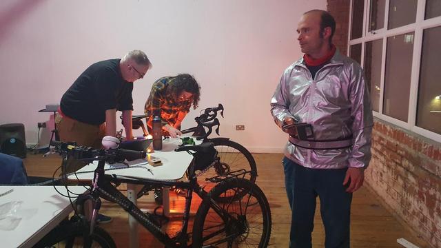 The the white DoES Liverpool Events space, Guy stands looking westward while in the back ground Stewart and Adrian look down at Stewart's bike. A gentle orange glow from Stew's My Bikes Git LED lights up their faces.