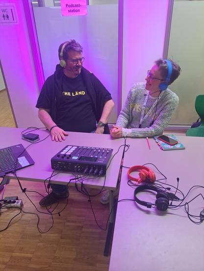 An einem Tisch mit Technik und Kopfhörer sitzen zwei Menschen und lachen sich gegenseitig an