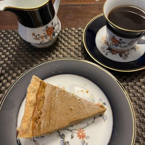 A slice of pumpkin pie on a white and blue china plate, alongside a matching cup of black coffee and a creamer.