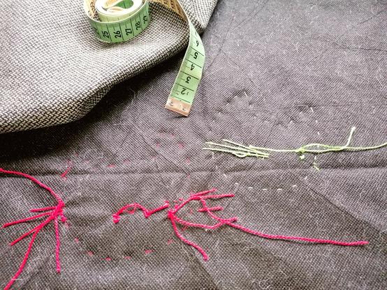 Black textile protector material with stitches and knots in green respectively red thread. Silhouettes with pointy ears and maybe parts of whisker-shaped thread. Chaotic. Backside of a piece of work. Woven fabric in grey-black and an ancient green and beige tape measure, almost completely rolled up lie there too