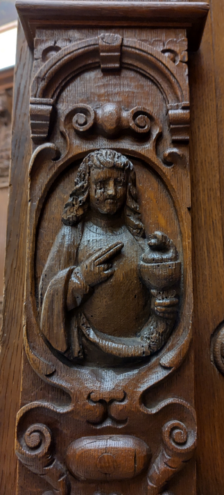 17th century oak carving. The image shows detail from a series of panels depicting biblical scenes. A man gestures with two fingers as if to shoot what looks like a monkey in a goblet.
