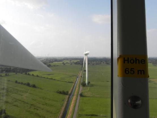 Von der Aussichtskabine eines begehbaren Windrads aus sind auf 65 Metern Höhe, wie ein Aufkleber an einer Stahlstrebe der Verglasung angibt, weitere Windräder, Felder und Wiesen sowie ein Stück eines gerade vorbeirotierenden Rotorblattes zu sehen.
