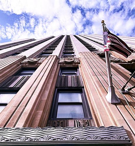 Color image of the Huntington Bank Tower, c. 1931, Art Deco, terra cotta and granite, 27 stories, Akron, Summit County, Ohio, USA. Image shows a view up the South Main Street front facing door, a flagpole on the right above the entry awning, a series of windows going up 27 floors, ending in a gauzy, cloudy bright blue sky.