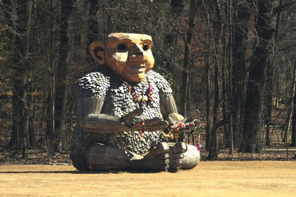 A very large wooden sculpture of a troll seated on a dry lawn with trees in the background. The troll's outstretched hands hold strings of colorful objects, and a necklace of colorful objects hangs across its chest.