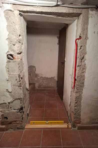 The image shows a narrow passage leading to a worn brown door with a rough, aged appearance. The walls are made of a mix of exposed bricks, crumbling plaster, and rough concrete, giving the space an old, unfinished look. The ceiling has visible cracks and a white electrical conduit running along it. The floor is covered with reddish-brown terracotta square tiles, contrasting with the rugged walls. A red crowbar leans against the right wall, suggesting ongoing work. A yellow spirit level rests on a wooden beam positioned across the entrance, possibly for measuring or alignment purposes. The space has a rustic, basement-like atmosphere, with signs of wear and renovation.
