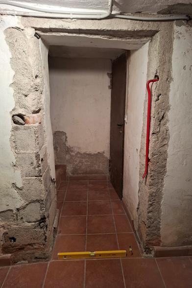 The image shows a narrow passage leading to a worn brown door with a rough, aged appearance. The walls are made of a mix of exposed bricks, crumbling plaster, and rough concrete, giving the space an old, unfinished look. The ceiling has visible cracks and a white electrical conduit running along it. The floor is covered with reddish-brown square tiles, contrasting with the rugged walls. A red crowbar leans against the right wall, suggesting ongoing work. A yellow spirit level rests on the floor positioned across the entrance, possibly for measuring or alignment purposes. The space has a rustic, basement-like atmosphere, with signs of wear and renovation.