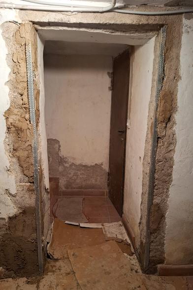 The image shows ongoing renovation work in a narrow passage with an aged brown door at the end. Two perforated metal strips have been installed vertically on both sidewalls, embedded in fresh plaster or mortar, likely for structural reinforcement or alignment. The floor is partially covered with protective cardboard sheets, suggesting active construction. A red marking on the tiled floor appears to be a reference line for further work. The rough, unfinished walls and visible repairs indicate that the space is being prepared for the next stage of restoration.
