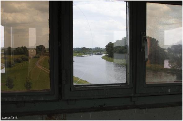 Blick von einem alten DDR-Grenzwachturm, eine Front mit 3 großen Fenstern, wobei das mittlere eine klare Sicht auf einen kleinen Sportboothafen freigibt.
Von rechts unten bis zum Hafen führt ein kleiner Seitenarm der Elbe. Längs dieses Arms sind Teile des Elberadwegs in Richtung Wittenberge zu sehen.
In den äußeren Scheiben spiegeln sich Teile der Umgebung wieder, links am Horizont der Rand von Lenzen, rechts schimmert zwischen Bäumen die Elbe durch.
Der Himmel ist stark bewölkt. Das Foto entstand bei Hochwasser.