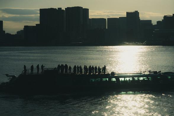 The image shows a silhouette of a boat with people standing on it, set against a city skyline. The scene is captured during sunset or sunrise, with the sun reflecting off the water, creating a dramatic and atmospheric effect. The city buildings are darkened in the background, providing a clear contrast to the brighter reflections on the water.