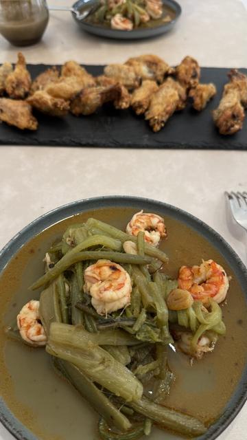 En la imagen se muestra un plato de comida casera servido sobre una mesa. En primer plano, hay un plato hondo con un guiso de verduras de hoja larga y camarones en un caldo de color marrón claro, con algunos trozos de ajo visibles. En segundo plano, sobre una bandeja negra de pizarra, hay alitas de pollo doradas y crujientes, dispuestas en fila. También se ve otro plato con el mismo guiso y un recipiente con una salsa espesa y oscura. La presentación es sencilla, y los platos parecen formar una combinación de sabores entre lo caldoso y lo crujiente.