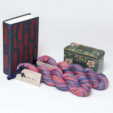 Picture shows two skeins of yarn positioned diagonally in shades of red and dark blue. At the head of the yarn stood on its end is a copy of the Penguin Clothbound Classic edition of Charles Dickens' A Tale of Two Cities. The book is dark blue with a repetitive red knitting pattern on it. Behind the yarn is a tin of Victorian style tea. The tin is green with an ornate floral pattern on it.