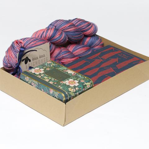 Picture shows an open A4 box contianing two skeins of yarn in shades of red and dark blue, a copy of Charles Dickens' A Tale of Two Cities and a tin of tea.