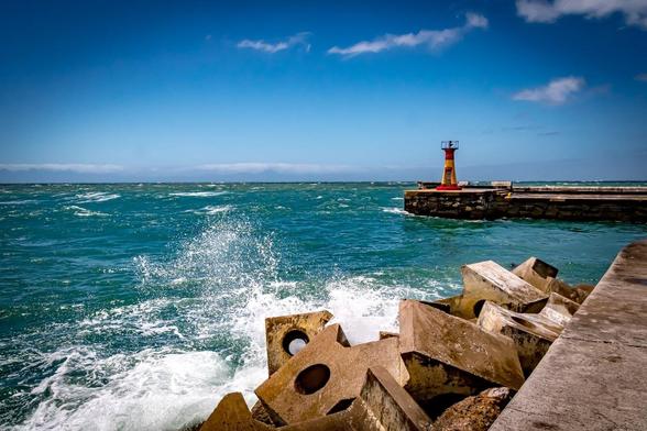 The image shows a coastal scene with waves crashing against large concrete wave breakers on the shore. In the background, there's a breakwater extending into the sea with a small, red-and-yellow lighthouse or navigational marker. The sky is clear with a few clouds.