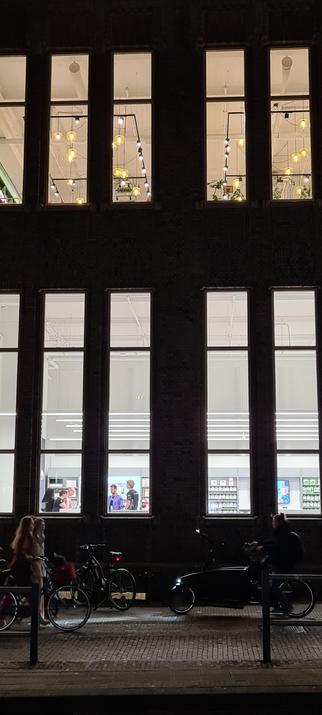 Photo of a two story, high ceilinged building at night. Both floors have narrow, extremely high windows. There are several people and display cases, lit by harsh white fluorescent light, visible through the ground floor windows. The second floor windows show the lightning rigs on the ceiling with various white and yellow spotlights.