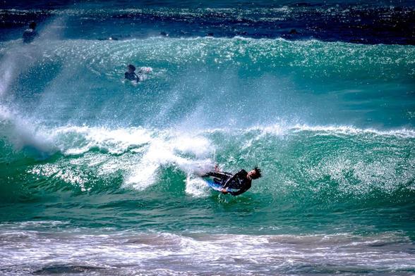 This image shows a person surfing on a wave in the ocean. The surfer is navigating through a breaking wave, dressed in a wetsuit and riding a surfboard. Other surfers can be seen in the background, waiting in the water. The wave is quite dynamic, with white water spray visible.