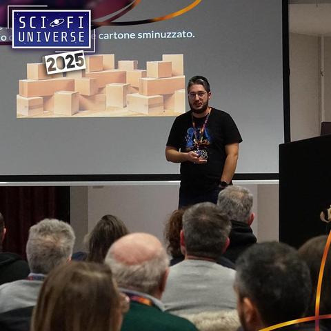 Un uomo con la barba e gli occhiali, indossa una maglietta nera con una stampa colorata, si trova su un palco di fronte a un pubblico. Alle sue spalle c'è uno schermo con la scritta "SCI FI UNIVERSE" e l'anno "2025", insieme a un'immagine di scatole di cartone.