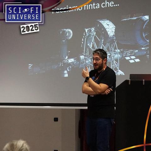 Un uomo con barba e occhiali è in piedi sul palco, con le braccia incrociate. Indossa una maglietta nera e jeans blu. Dietro di lui c'è uno schermo di proiezione che mostra un'immagine di una stazione spaziale e il testo "Facciamo finta che..." nell'angolo in alto a destra. Nell'angolo in alto a sinistra dello schermo di proiezione c'è un logo con la scritta "SCI-FI UNIVERSE" e "2025".