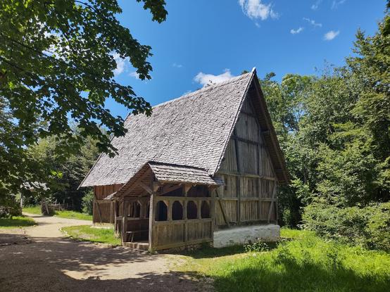 Das Bild zeigt ein traditionelles, historisches Fachwerkhaus mit einem steilen, mit Holzschindeln gedeckten Dach. Die Wände bestehen aus einer Kombination von Holzbalken und weiß verputztem Mauerwerk im unteren Bereich. An der Vorderseite des Gebäudes befindet sich ein kleiner, überdachter Vorbau mit geschnitzten Holzverzierungen und einer Sitzbank, der vermutlich als Eingangsbereich dient. Der Weg vor dem Haus ist unbefestigt und schlängelt sich durch die grüne Umgebung. Das Haus ist von dichtem Wald und üppiger Vegetation umgeben, wodurch eine idyllische, abgeschiedene Atmosphäre entsteht. Der Himmel ist strahlend blau mit einigen kleinen Wolken, und das Sonnenlicht wirft sanfte Schatten auf den Boden, während die Blätter der Bäume im Vordergrund das Bild teilweise einrahmen.