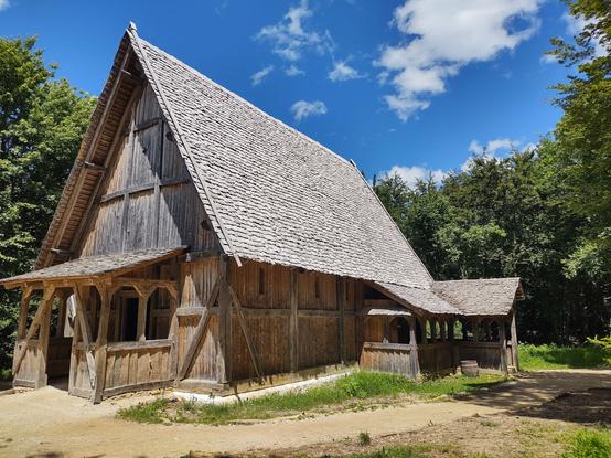 Das Bild zeigt ein großes, historisches Fachwerkhaus mit einem sehr steilen Dach, das mit Holzschindeln gedeckt ist. Die Wände bestehen aus groben Holzbalken, die das traditionelle Fachwerk betonen. Der Eingang des Hauses verfügt über ein kleines, überdachtes Vordach, das auf stabilen Holzbalken ruht. An der rechten Seite des Gebäudes ist eine überdachte, offene Galerie mit Holzgeländer zu sehen, die sich entlang der Hauswand erstreckt. Der Boden besteht aus einem unbefestigten Weg, der sich harmonisch in die natürliche Umgebung einfügt. Das Haus ist von dichtem, grünem Wald umgeben, der einen starken Kontrast zum strahlend blauen Himmel mit vereinzelten weißen Wolken bildet. Die Szene wirkt ruhig und idyllisch, als wäre das Gebäude Teil eines Freilichtmuseums oder einer historischen Rekonstruktion.