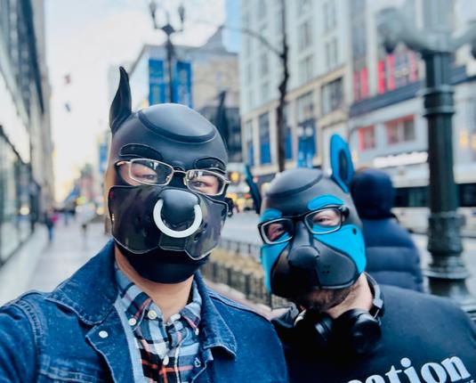 This image places our pup explorers within Chicago's vibrant streetscape. The black/camo hood with its striking white ring and camouflage muzzle complement the wearer's casual denim jacket and plaid button-up, while the blue-accented hood partner appears with subtle headphones around their neck. The urban composition captures the soft glow of early evening light washing across the downtown buildings, with street lamps beginning to illuminate against the blue-gray sky.
The contrast between the precisely crafted neoprene hoods and the everyday urban environment creates a compelling visual narrative about identity expression within public spaces. Their confident posture and direct gaze suggest comfortable self-possession amidst the city's flow, while the slightly defocused background hints at the energy of Chicago's downtown core.