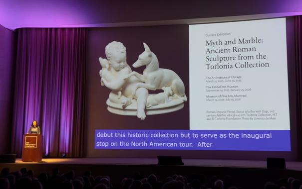 The image captures the sophisticated ambiance of the Art Institute of Chicago's auditorium bathed in rich purple lighting. Presenter Lisa Ayla Çakmak stands at the wooden podium while the projection screen displays the exhibition title "Myth and Marble: Ancient Roman Sculpture from the Torlonia Collection" in an elegant typography designed specifically for the exhibition. The centerpiece of the slide features a hauntingly beautiful 2nd-century marble sculpture of a young boy with dogs, creating a poignant visual dialogue between ancient and contemporary interpretations of human-canine connections. The warm marble tones contrast dramatically against the deep burgundy background, while exhibition dates reveal this as a prestigious North American tour running from March 15 to June 29, 2025.
The delicate craftsmanship of the ancient Roman sculpture is evident even in projection form, with the smooth marble surface capturing light in a way that brings dimension to the child's serene expression and the attentive posture of the canine figure beside him. The exhibition information suggests an exceptional cultural moment - these treasures have never left Europe, making their Chicago debut particularly significant.