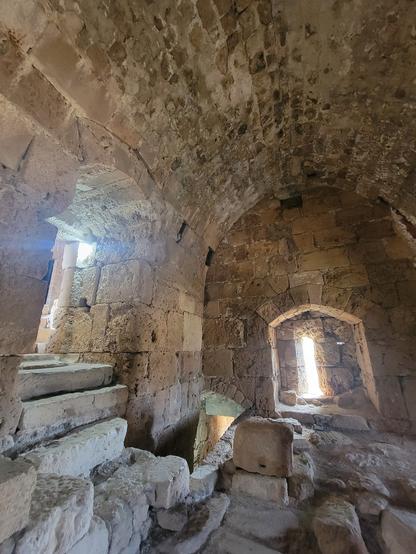 Inside a small room in a stone castle. The ceiling is arched, and the wall at the far end of the room has a very narrow window letting in the bright light from outside. There is also a small window or exit to the left.