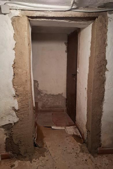 The image shows ongoing renovation work in a narrow passage with an aged brown door at the end. Two perforated metal strips have been installed vertically on both sidewalls, embedded in fresh mortar. The floor is partially covered with protective cardboard sheets, suggesting active construction. The rough, unfinished walls and visible repairs indicate that the space is being prepared for the next stage of restoration.