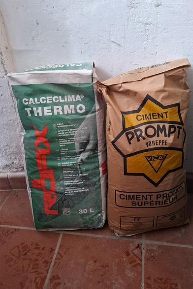 The image shows two natural mortar bags on a terracotta floor with a white plastered wall in the background. The left bag is a special natural thermo-insulating mortar in a green package and the right one is a natural quick-setting cement in a yellow-brown package.