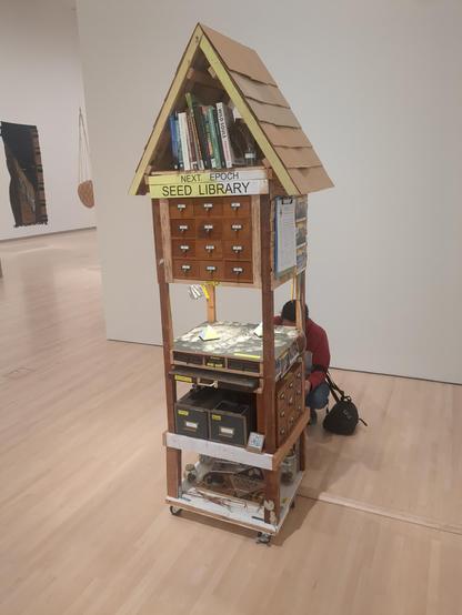 Picture of a six foot tall wooden structure in an art gallery. The structure is roughly 2x2x6 ft. The top is a pitched roof with books about plants and nature in it, below that is a sign that says "Next Epoch Seed Library", below that is a series of 12 drawers (in rows of 3) labeled alphabetically. Below that two ceramic pyramids sit on a platform with overhead lighting. Below that are a few more labeled drawers, and a the bottom is a platform with various pieces of plants.