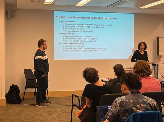 Two people presenting at a conference. Several audience members are visible from behind.